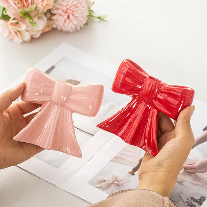 Hands hold pink and red ceramic bow candle holders against a white background.