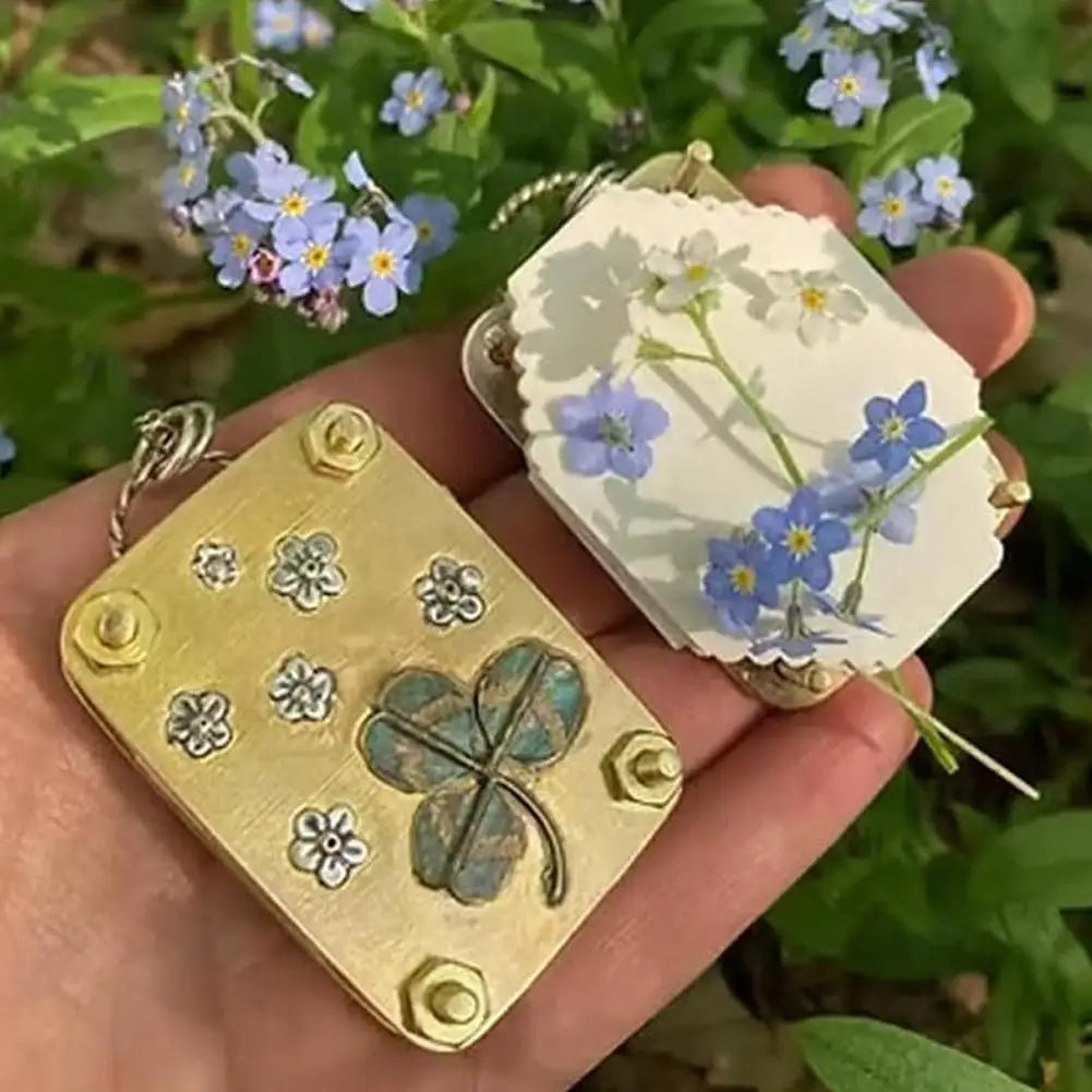 A hand holds a gold-colored metal flower press kit with a pressed blue flower and a white piece of dried flower.