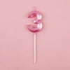 A pink number three candle with a thin stick against a light pink background.