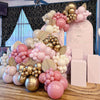 A pink and gold balloon garland arch display featuring various sized balloons and confetti on a wooden surface.
