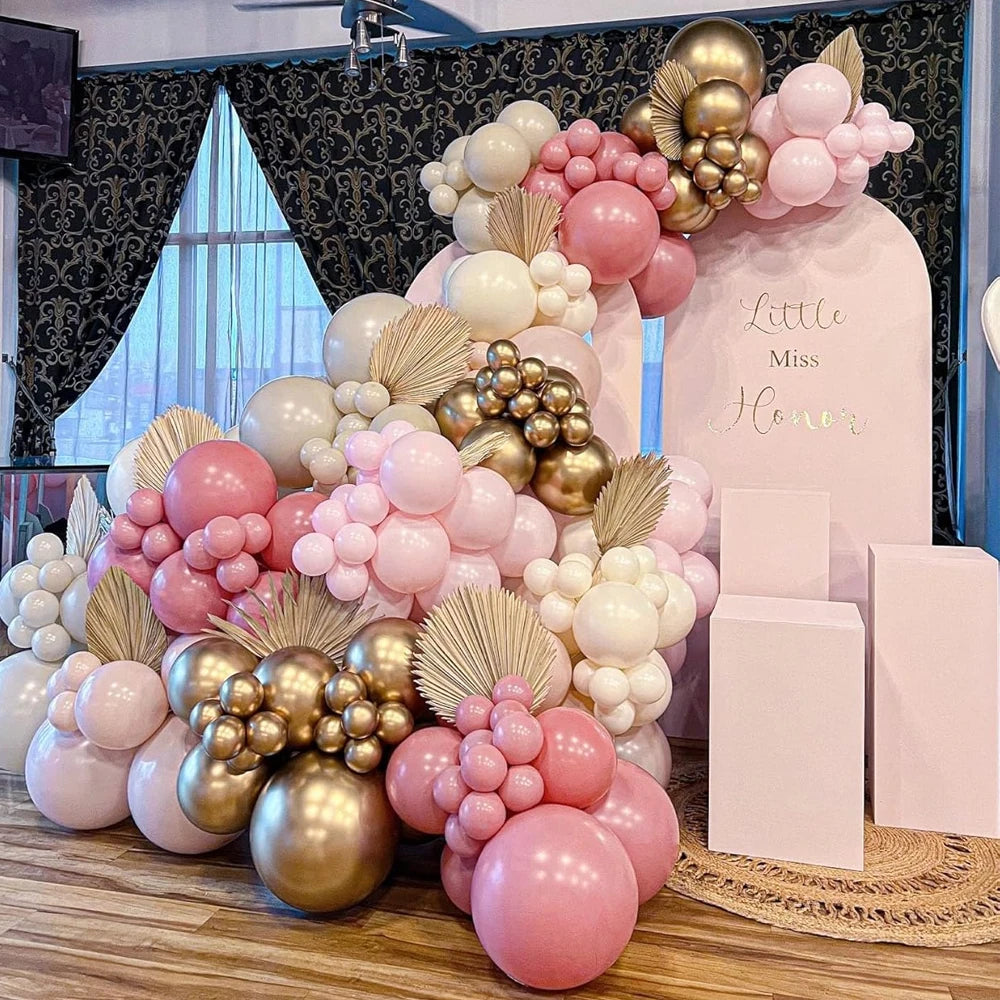 A pink and gold balloon garland arch display featuring various sized balloons and confetti on a wooden surface.