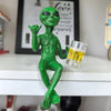 A green resin alien statue with an outstretched hand holding a glass of beer on a white surface.
