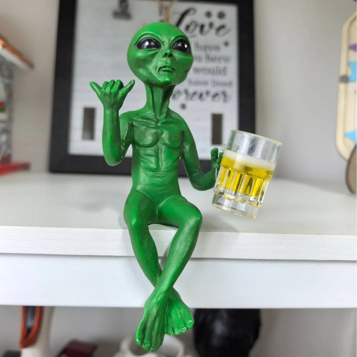 A green resin alien statue with an outstretched hand holding a glass of beer on a white surface.