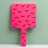 A pink handheld vanity mirror with an ergonomic handle featuring a pattern of black eyelashes against a light green backgroun