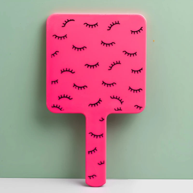 A pink handheld vanity mirror with an ergonomic handle featuring a pattern of black eyelashes against a light green backgroun