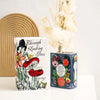 A white ceramic book-shaped vase with floral designs sits next to dried flowers.