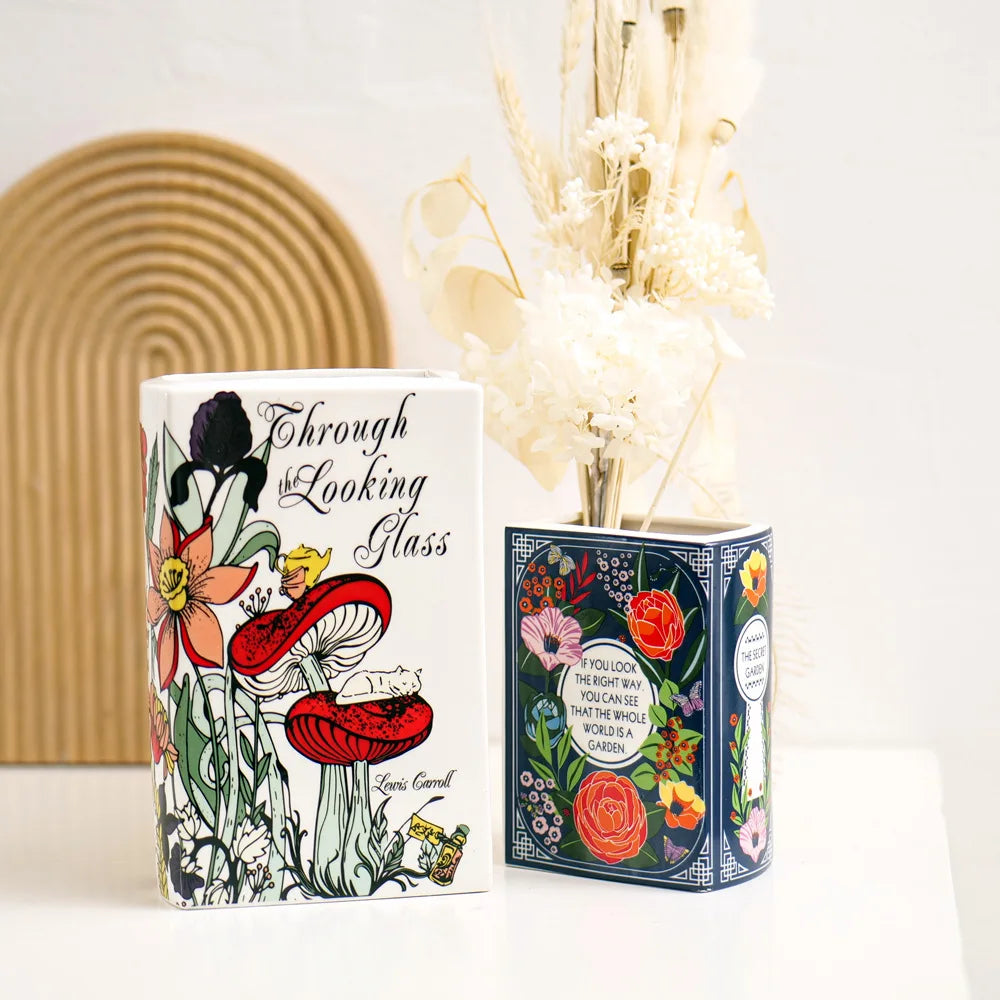A white ceramic book-shaped vase with floral designs sits next to dried flowers.
