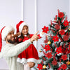 A man and a girl in festive attire are holding a decorated Christmas tree with red flowers.