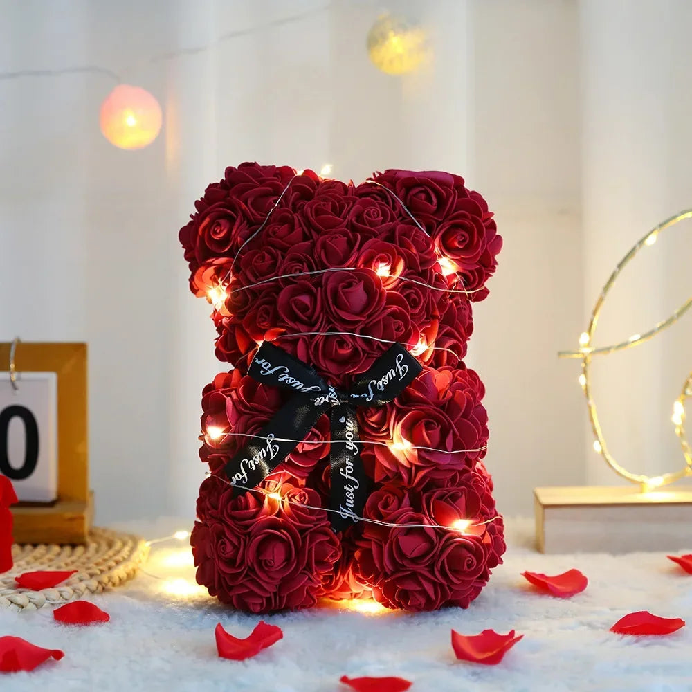 A dense red artificial rose teddy bear illuminated by warm string lights on a white surface.
