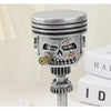 A silver mechanical piston skull sculpture with gears and a cylindrical top, displayed on a white surface.