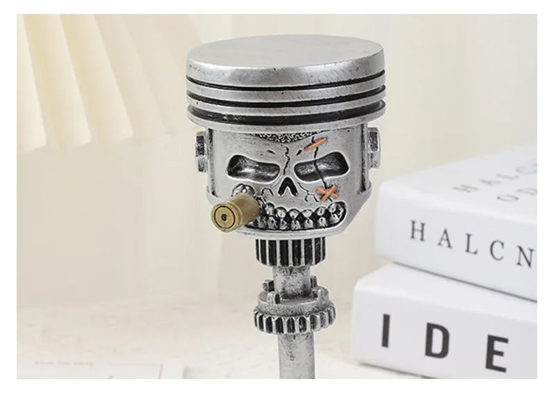 A silver mechanical piston skull sculpture with gears and a cylindrical top, displayed on a white surface.
