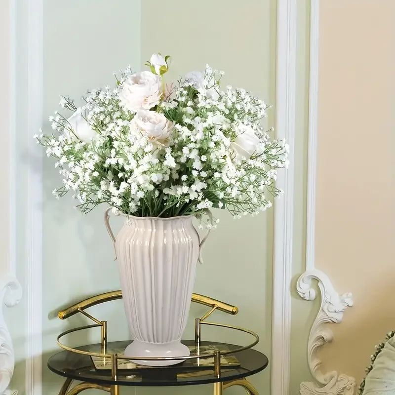 A vase filled with white baby's breath artificial flowers displayed on a gold-toned table against a pale green wall.