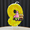 A yellow cake candle shaped like the number 0, decorated with glitter and featuring SpongeBob and Patrick characters.