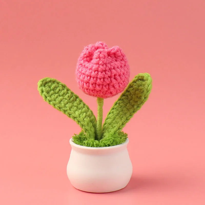 A hand-woven crochet pink tulip flower potted in a white ceramic container against a soft pink background.