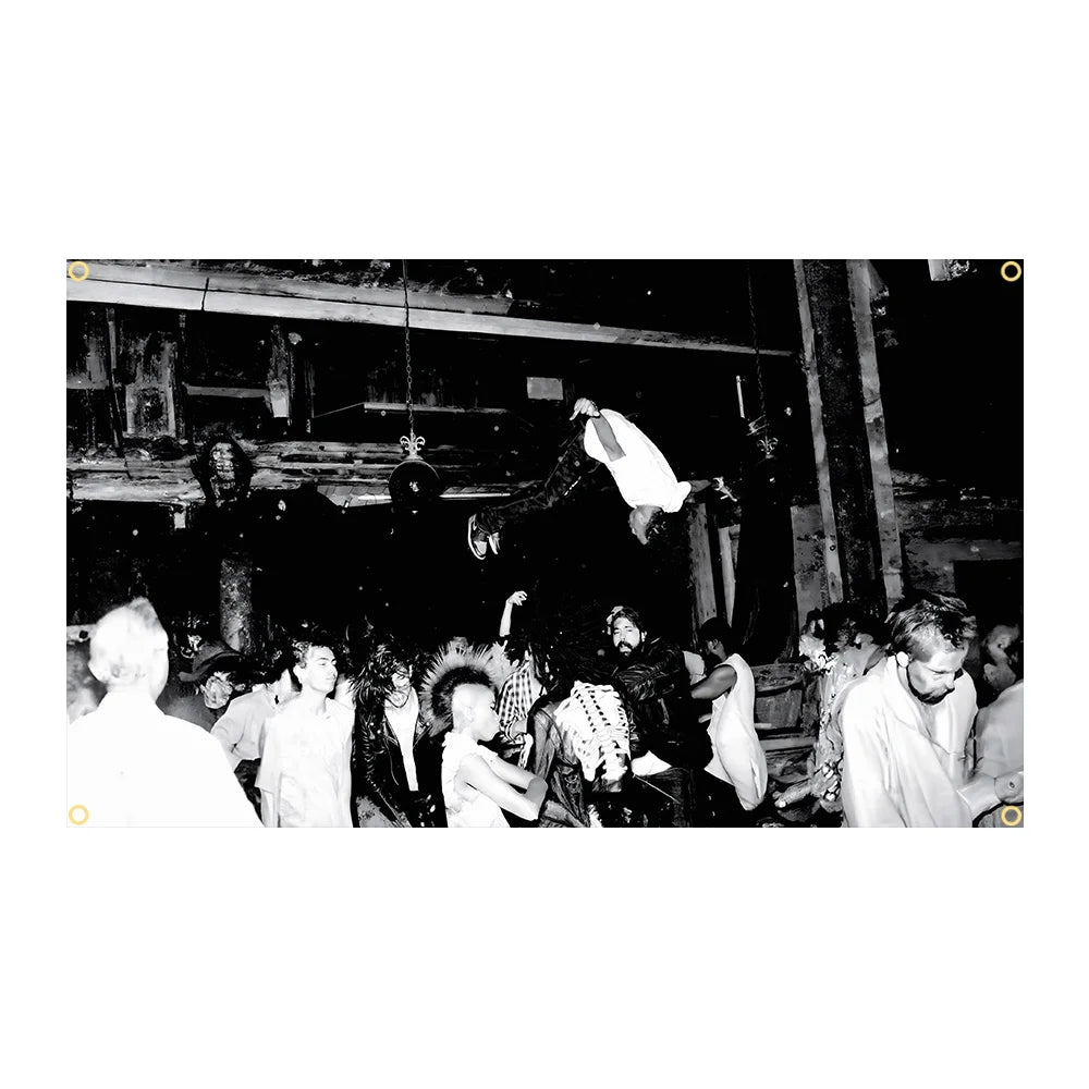 A black and white photograph of a group of people gathered in a dimly lit interior, with a person raising a white garment abo