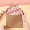 A hand is holding a brown envelope with a pink birthday cake design on the front.