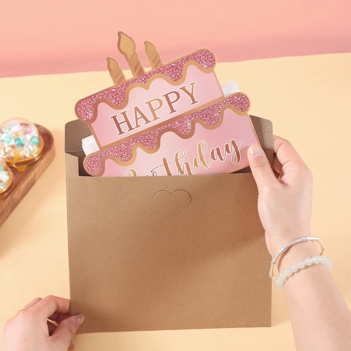 A hand is holding a brown envelope with a pink birthday cake design on the front.