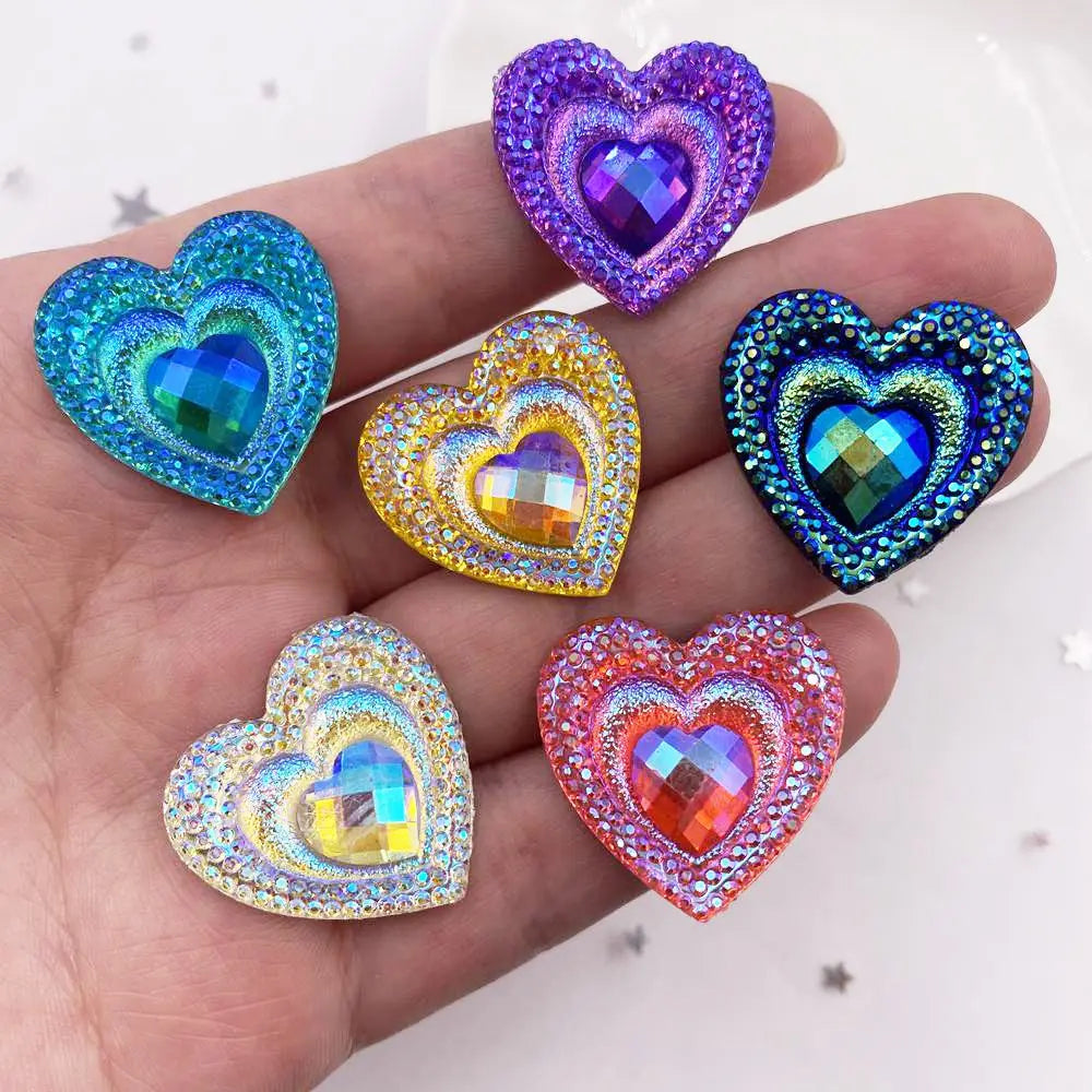 A hand holds a set of twelve heart-shaped iridescent blue and purple resin rhinestone flatbacks.