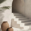 A terracotta vase sits on a white staircase in a bright, minimalist interior with palm leaves casting shadows.