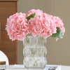 A glass vase filled with pink artificial hydrangea flowers sits on a white table next to a stack of books.