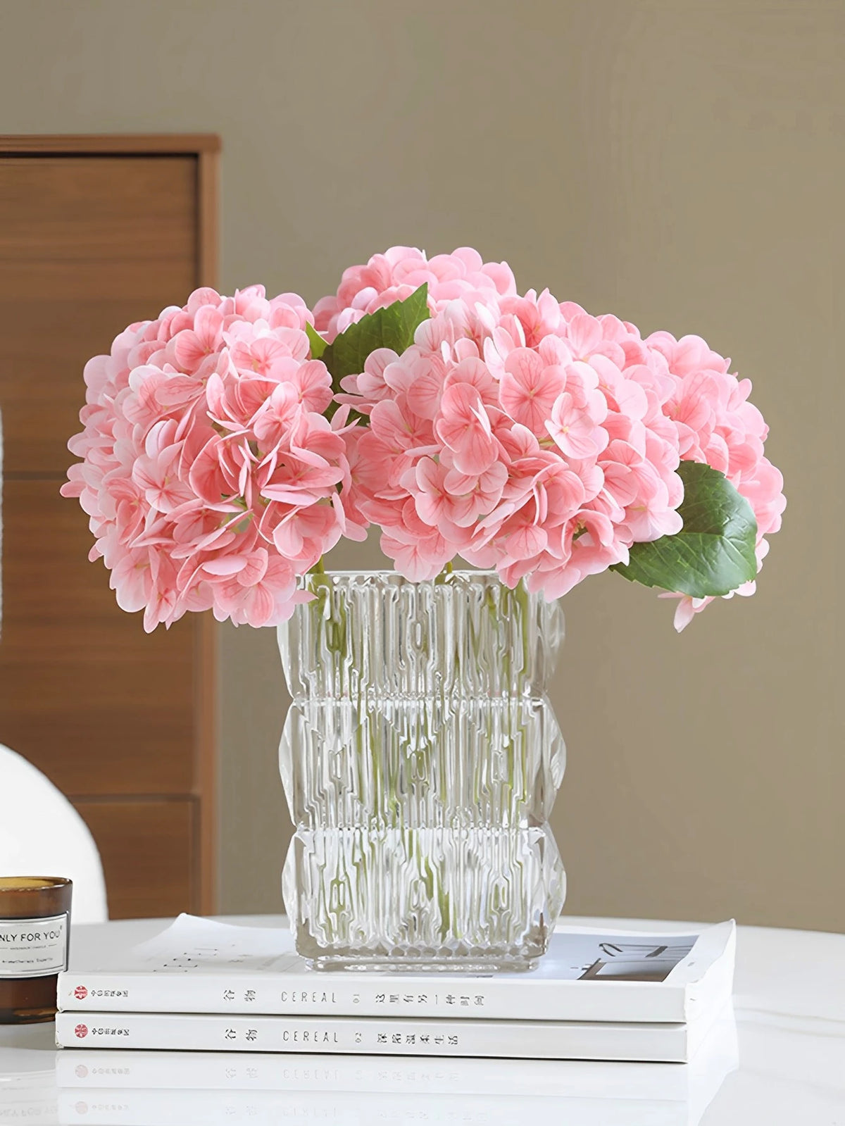 A glass vase filled with pink artificial hydrangea flowers sits on a white table next to a stack of books.