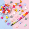 A collection of colorful silicone heart beads arranged in a circle and scattered on pink and blue surfaces next to a pen.