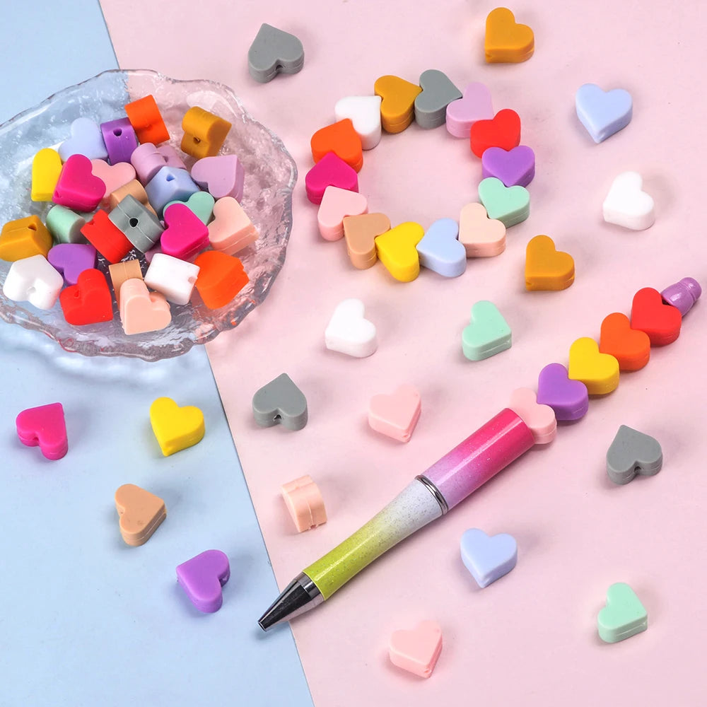 A collection of colorful silicone heart beads arranged in a circle and scattered on pink and blue surfaces next to a pen.
