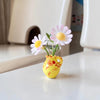 A yellow ceramic vase holding three pink and white daisy-like flowers on a white surface.