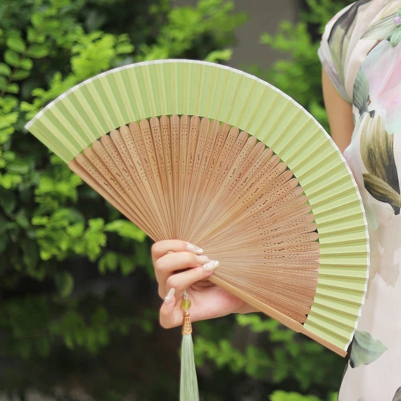 A person holds an elegant bamboo folding fan with a light green and natural wood finish against a blurred green outdoor backg