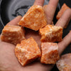 Several orange-colored natural raw healing crystal stones are held in a person's hand against a dark background.