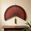 A handwoven bamboo wall fan decor with a reddish-brown color and a gold tassel mounted on a light wall.