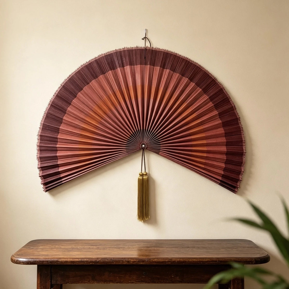 A handwoven bamboo wall fan decor with a reddish-brown color and a gold tassel mounted on a light wall.