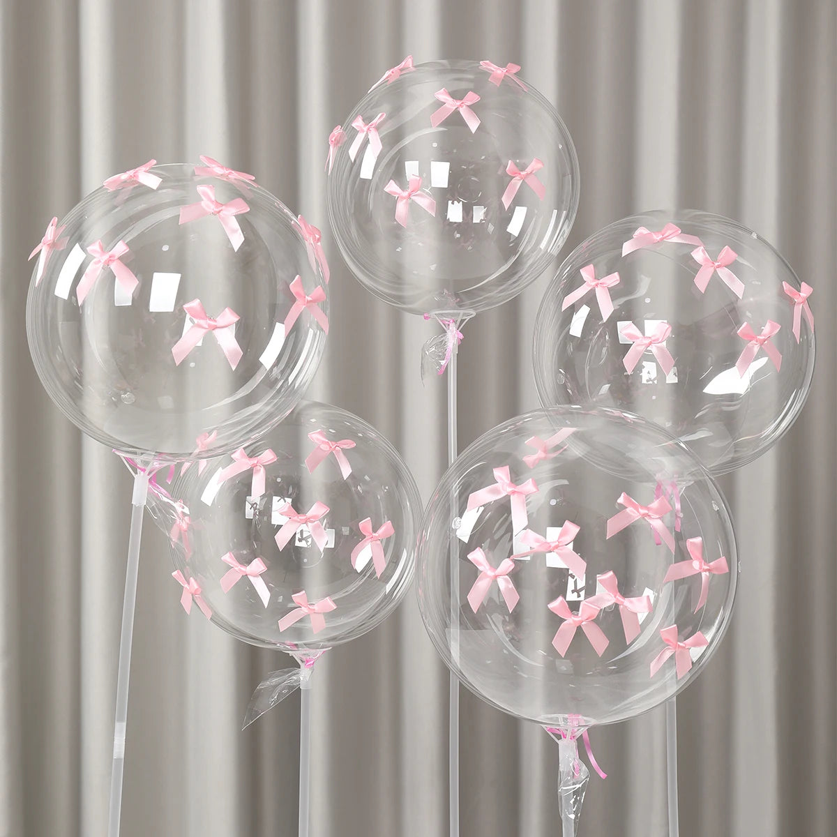 A set of clear latex balloons decorated with pink ribbon bows, displayed against a light gray curtain background.