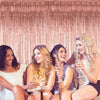 Four women smiling while holding champagne flutes in front of a shimmering rose gold curtain backdrop.