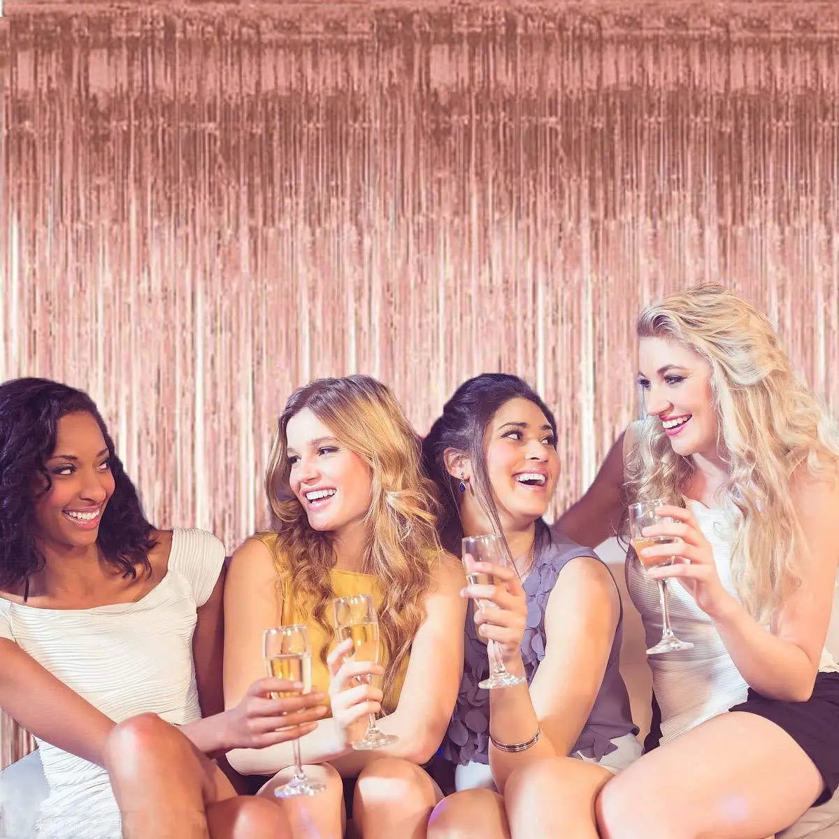 Four women smiling while holding champagne flutes in front of a shimmering rose gold curtain backdrop.