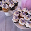 A platter of cupcakes decorated with white frosting and purple butterfly toppers on a white table.