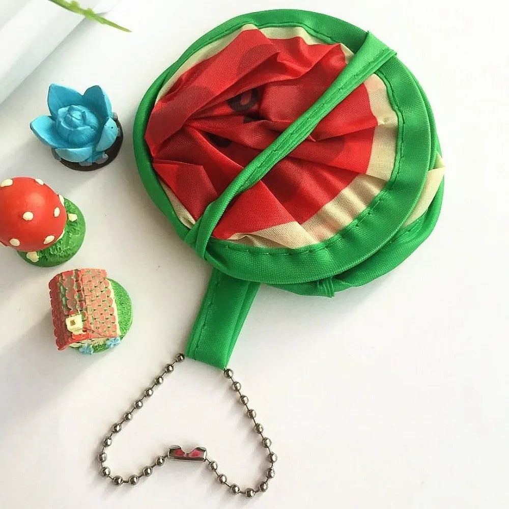 A green foldable hand fan with a red fruit design, next to a blue flower and a red and green ladybug.