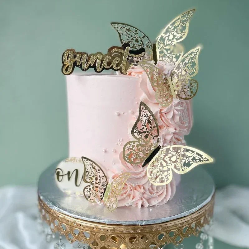 A pink buttercream cake decorated with rose petals and gold butterfly toppers spelling 'sweet' and 'one'.