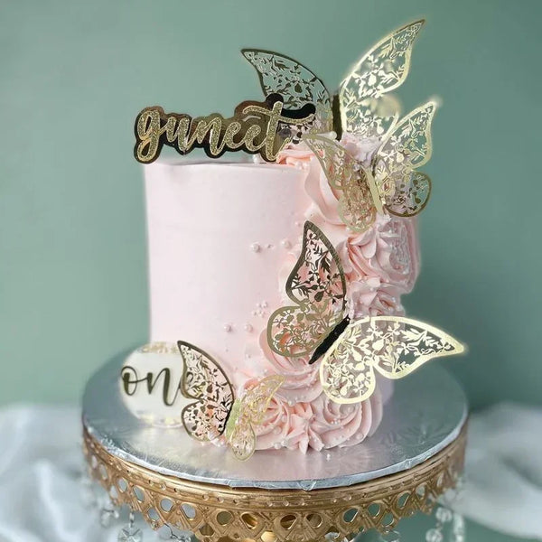 Pastel de crema rosa decorado con pétalos de rosa y toppers de mariposa dorada que deletrean 'sweet' y 'one'.