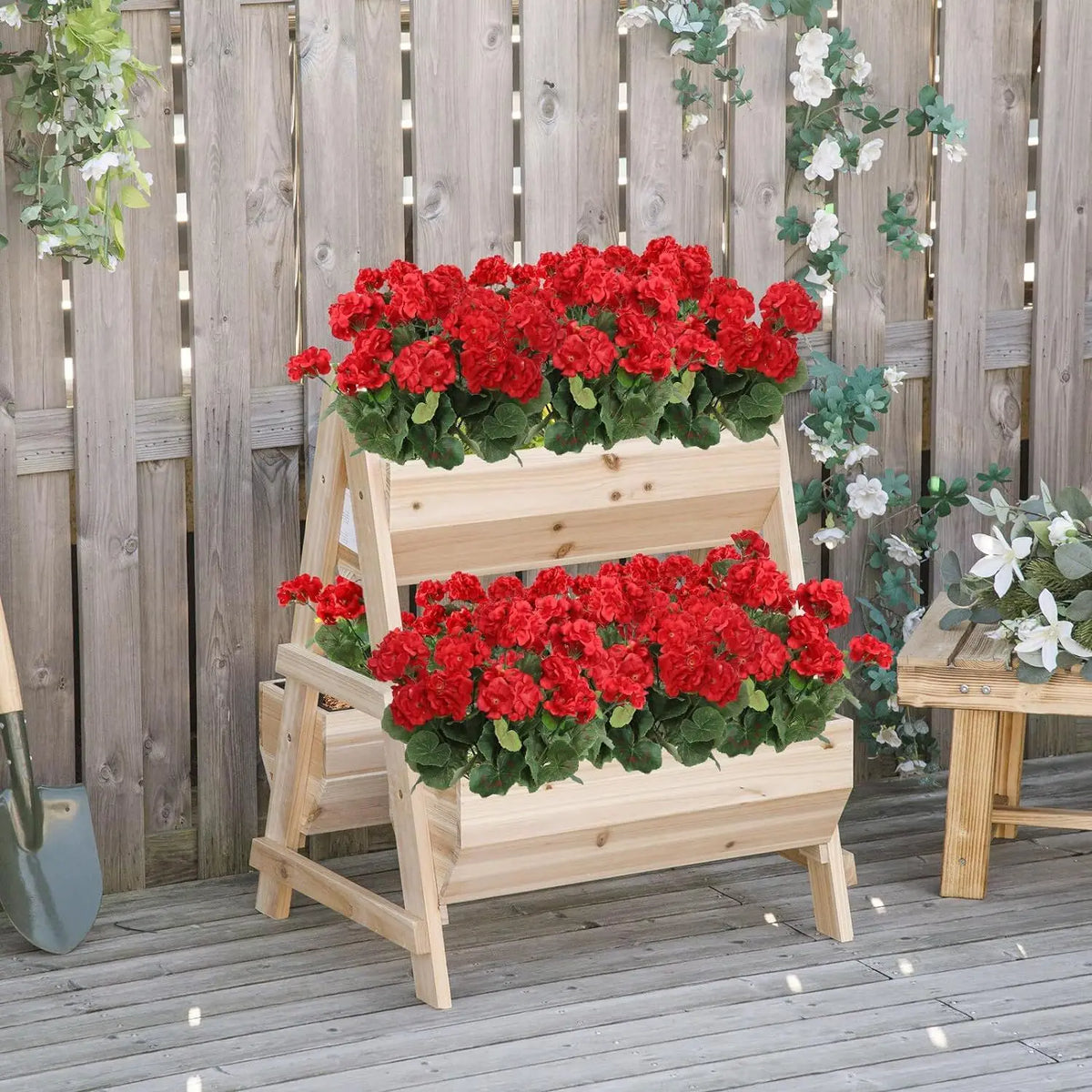 A wooden planter box filled with bright red artificial flowers set against a gray wooden fence.