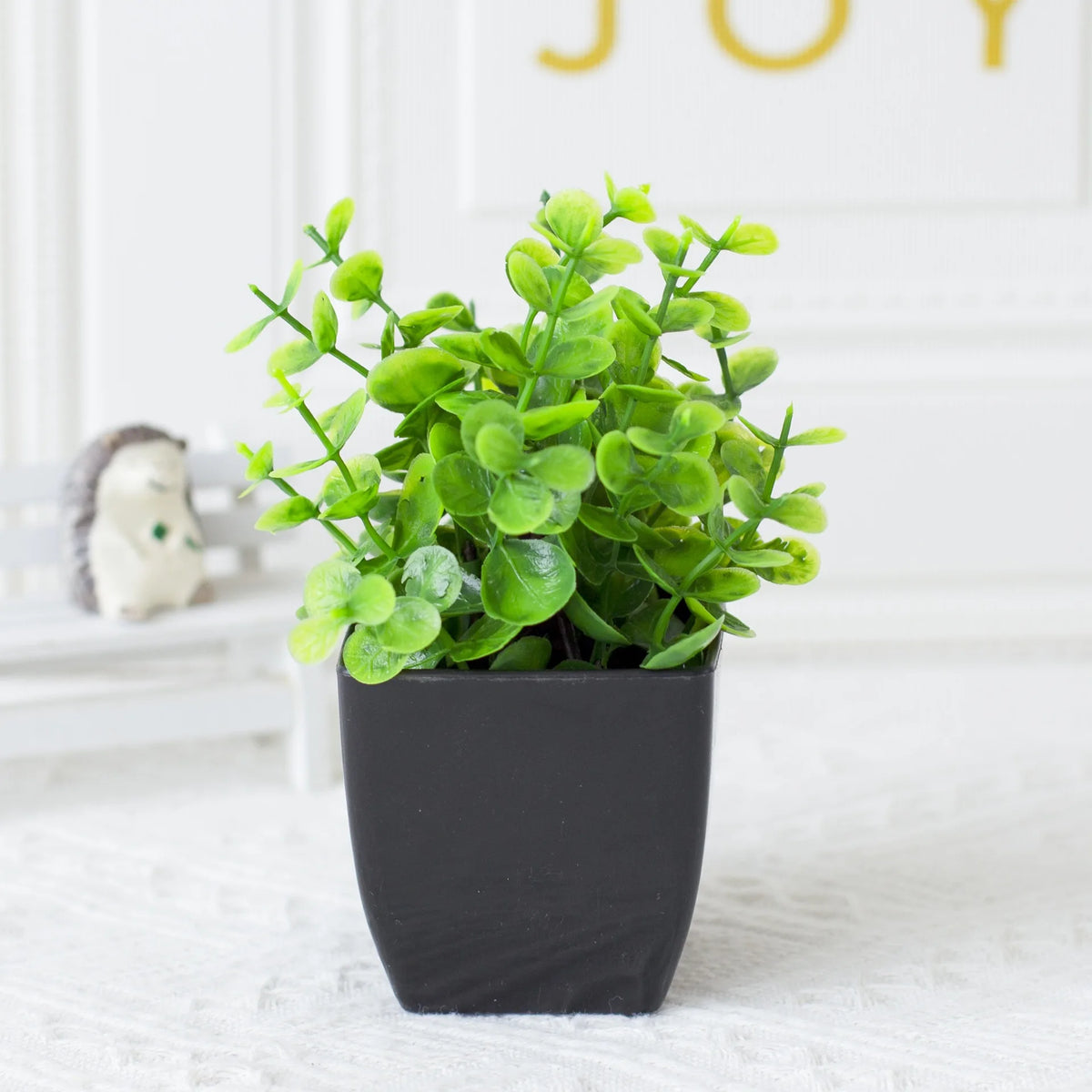 A small potted artificial eucalyptus plant in a black pot sitting on a white surface.