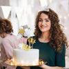 A smiling woman holds a white cake decorated with gold and silver crown toppers in a bright indoor setting.