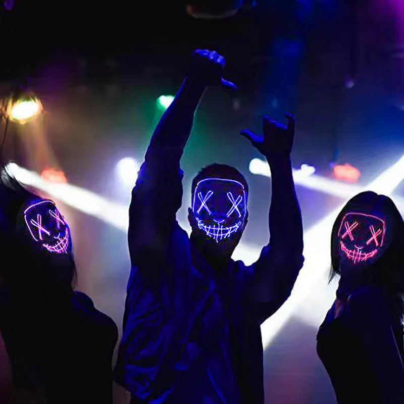 A person wearing a glow-in-the-dark LED purge mask with neon wire details raises their arms in a performance setting.