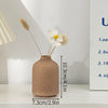 A minimalist ceramic vase in a beige tone holds dried floral elements on a white surface.