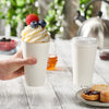 A hand holds a white disposable paper coffee cup filled with whipped cream and berries on a wooden table.