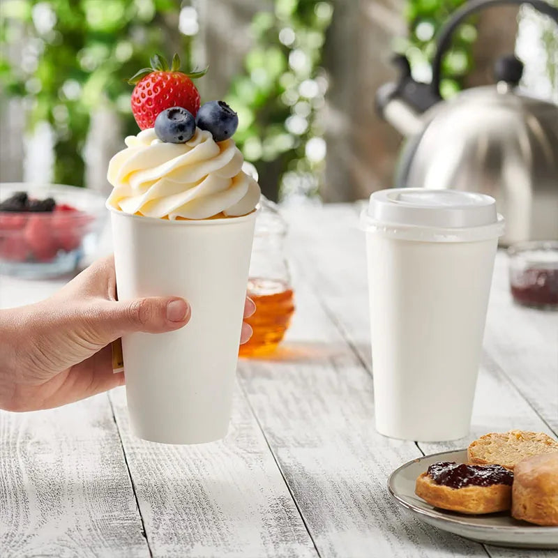 A hand holds a white disposable paper coffee cup filled with whipped cream and berries on a wooden table.