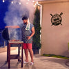 A man grilling outdoors with smoke, in front of a wall sign featuring a BBQ zone logo.
