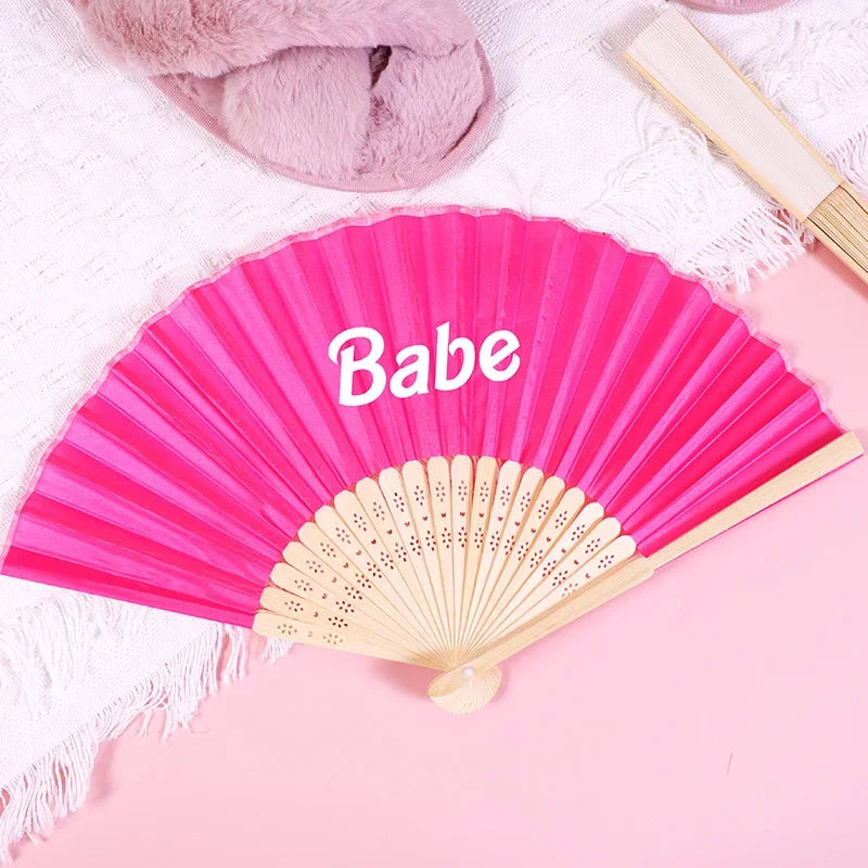 Pink hand fan with the name Babe written on it, resting on a pink surface.