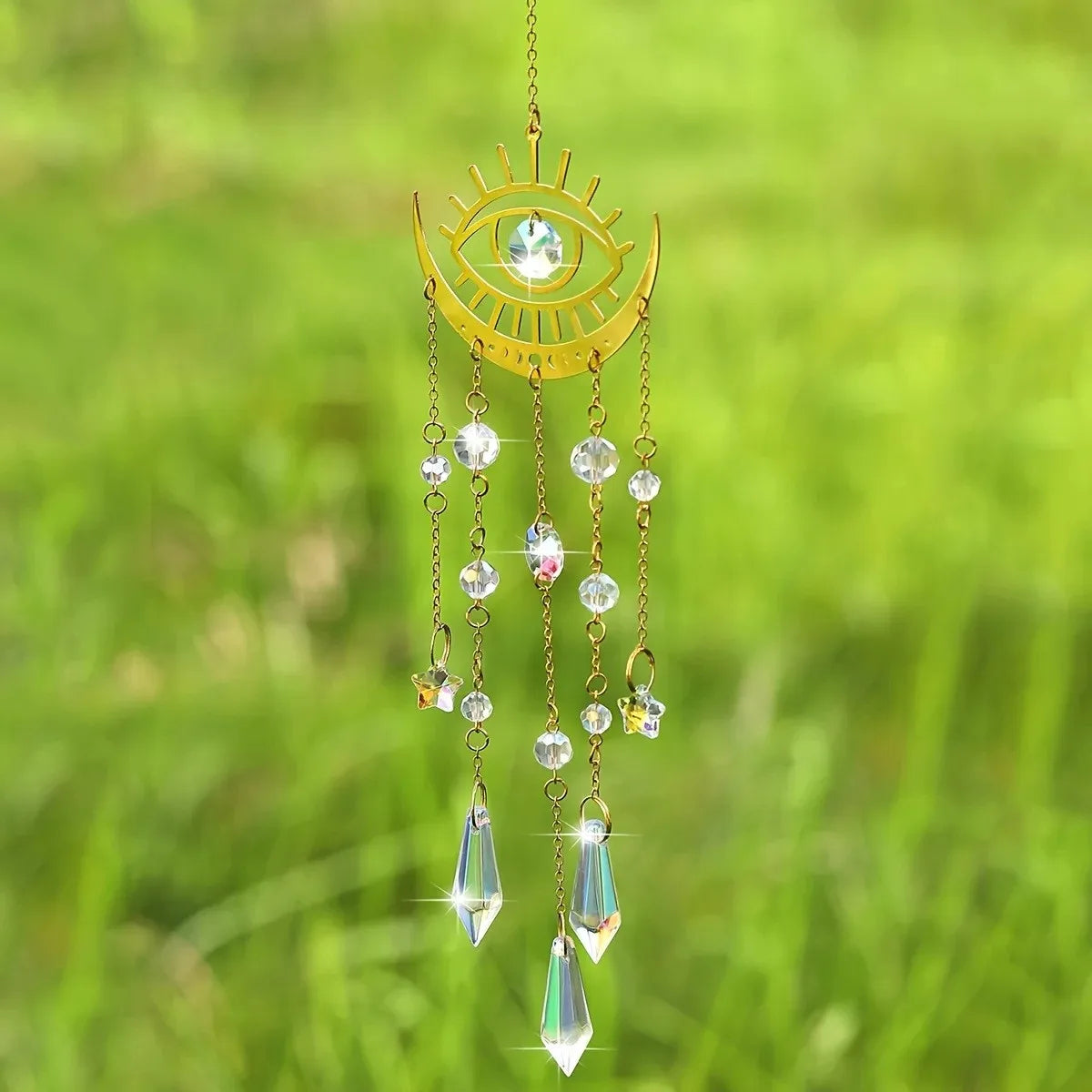 A gold flame moon crystal sun catcher with a central eye design and dangling crystals suspended against a green background.