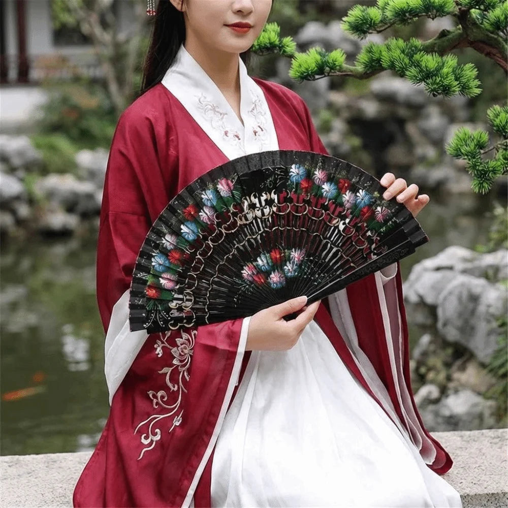 A woman in a red and white traditional dress holds a black decorative folding fan against a natural outdoor background.
